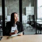 Portrait of a woman in a modern office pondering ideas, holding a notebook and pen.